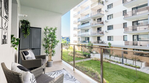 Een balkon met rieten meubilair, een bloempot en uitzicht op een meerlaags gebouw met balkons.