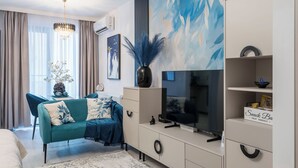 Een moderne woonkamer met een blauwgroene bank, een grote flatscreen televisie, een decoratieve vaas en een luchtverkoeler aan de muur.