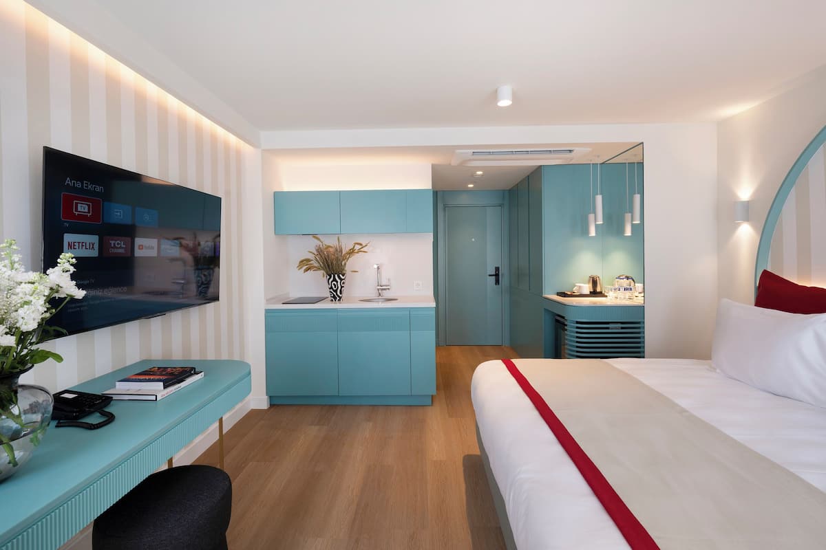 A modern hotel room with a TV, a small kitchenette, a bed, and a desk with a vase of flowers.