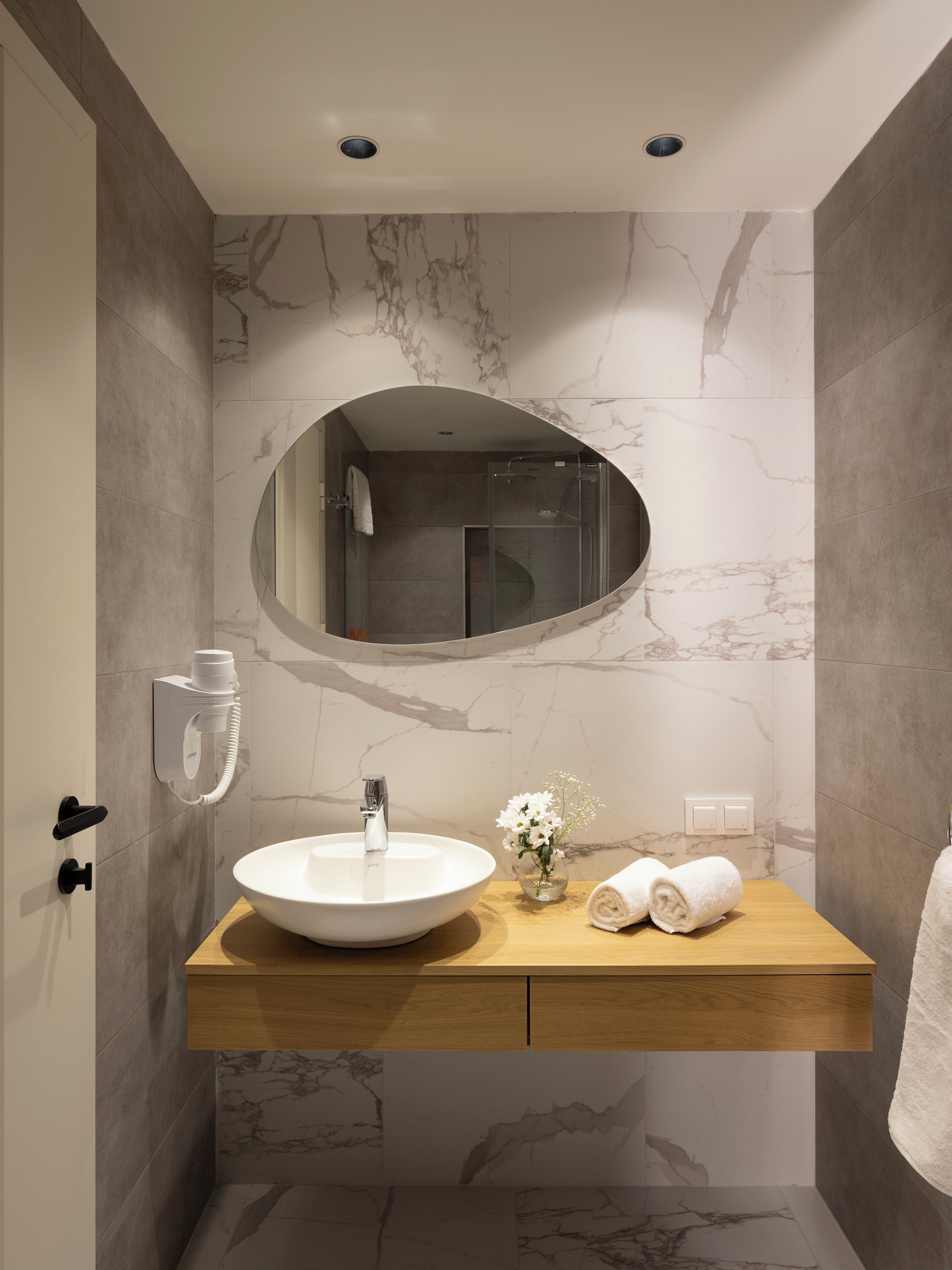 A modern bathroom with a marble wall, a round mirror, a white sink, and a wooden vanity.