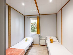 A small, single-room accommodation with two single beds, a window with yellow curtains, a radiator, and a view of greenery outside. - Cozy chalet for 6 with terrace and garden furniture (Badefols-sur-Dordogne)