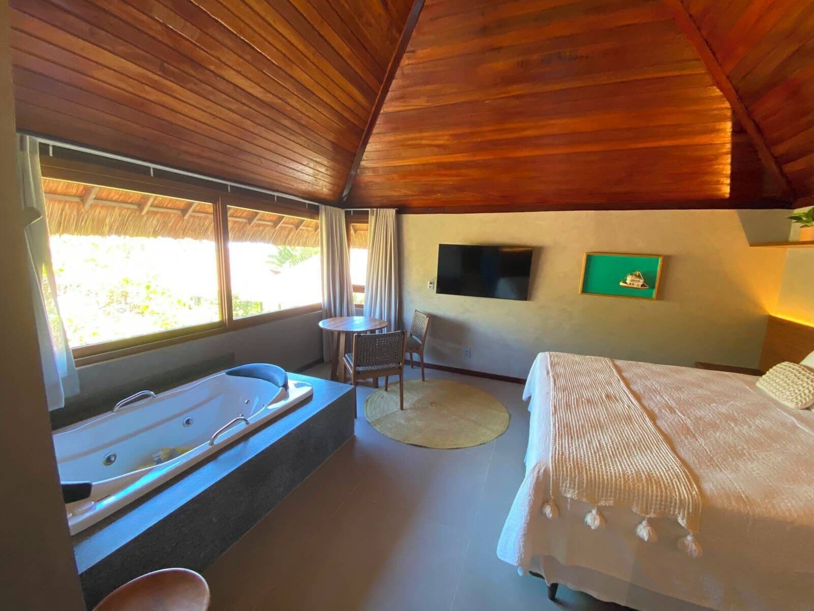 A room with a wooden ceiling, a large window, a bathtub, a bed, a chair, and a television.