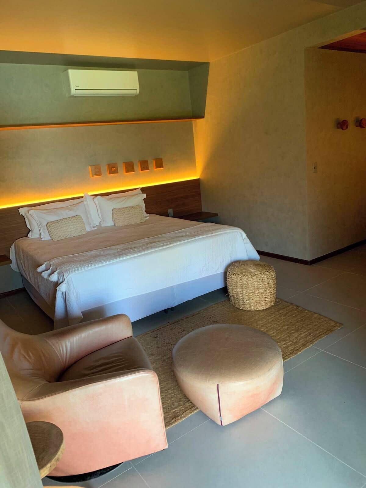 Un dormitorio moderno con una cama grande, cabecera con iluminación integrada, mesita de noche y un rincón de descanso con una silla y reposapiés.
