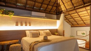 Een slaapkamer met een groot bed, een houten hoofdeinde, een klein tafeltje, een geweven kruk, een badkuip en een plafondventilator.