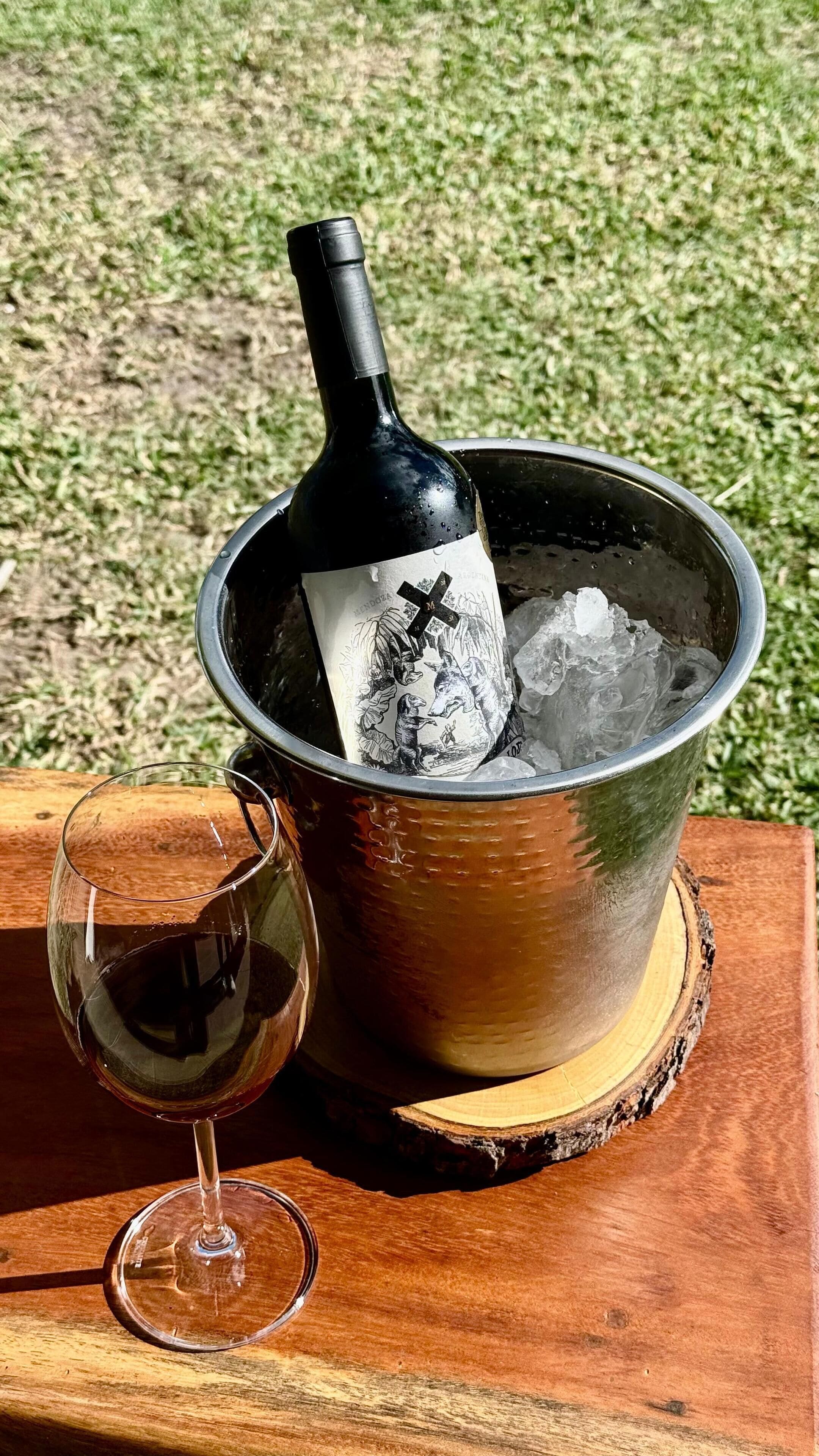 Una botella de vino en un balde de metal con hielo, una copa de vino y una superficie de madera.