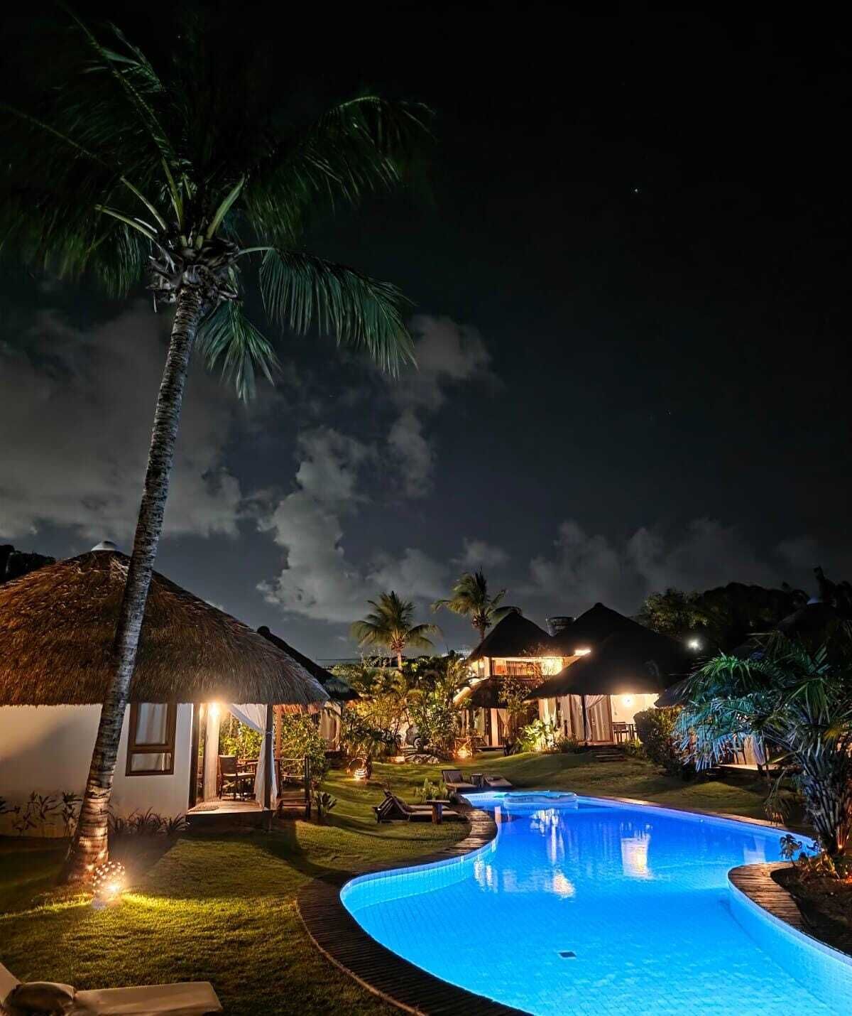 Un complexe hôtelier ouvert la nuit, doté d’une piscine, de bungalows aux toits de chaume et de palmiers.