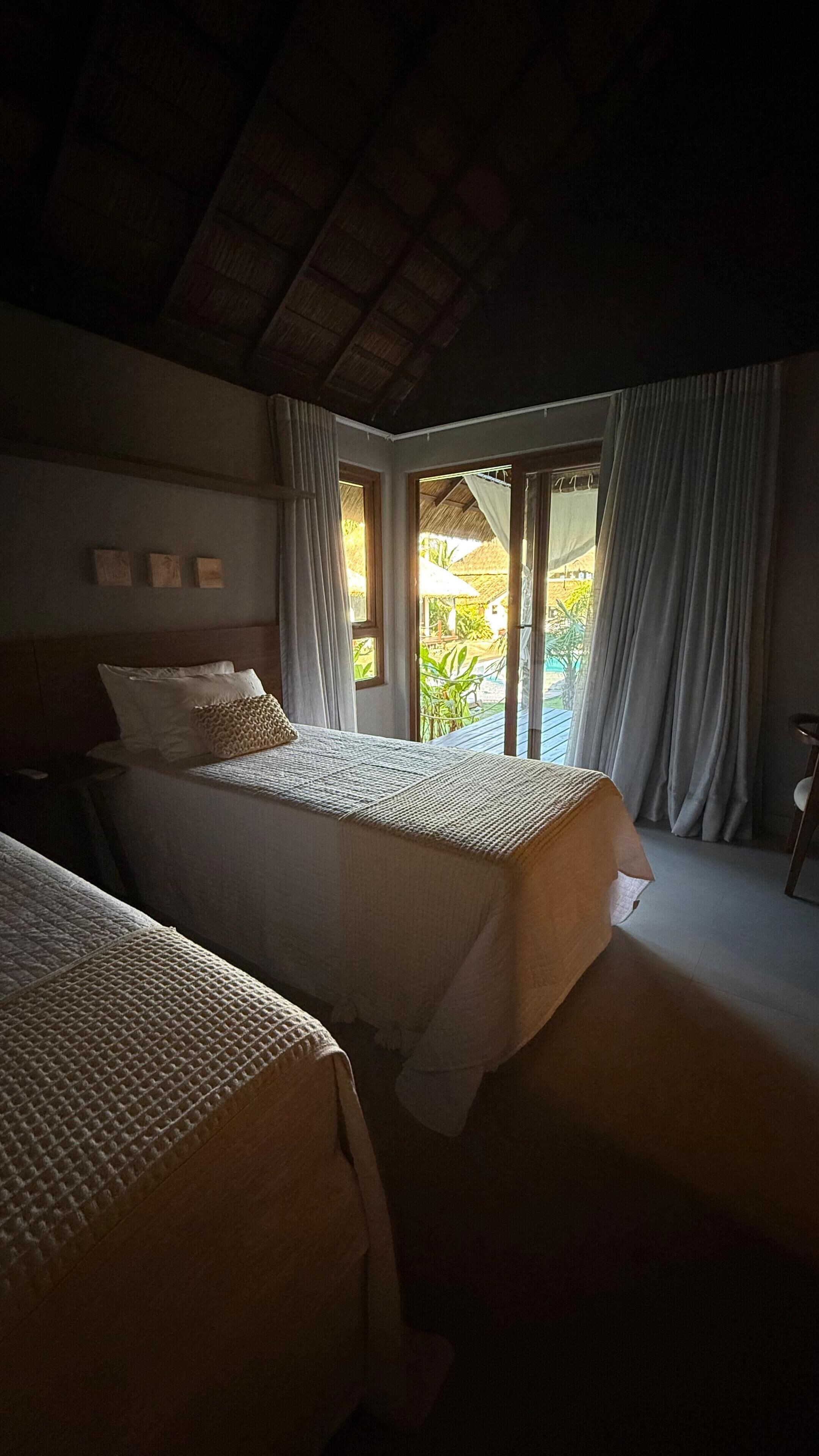 Une chambre avec deux lits, un plafond en bois et une vue sur un jardin grâce à de grandes fenêtres.