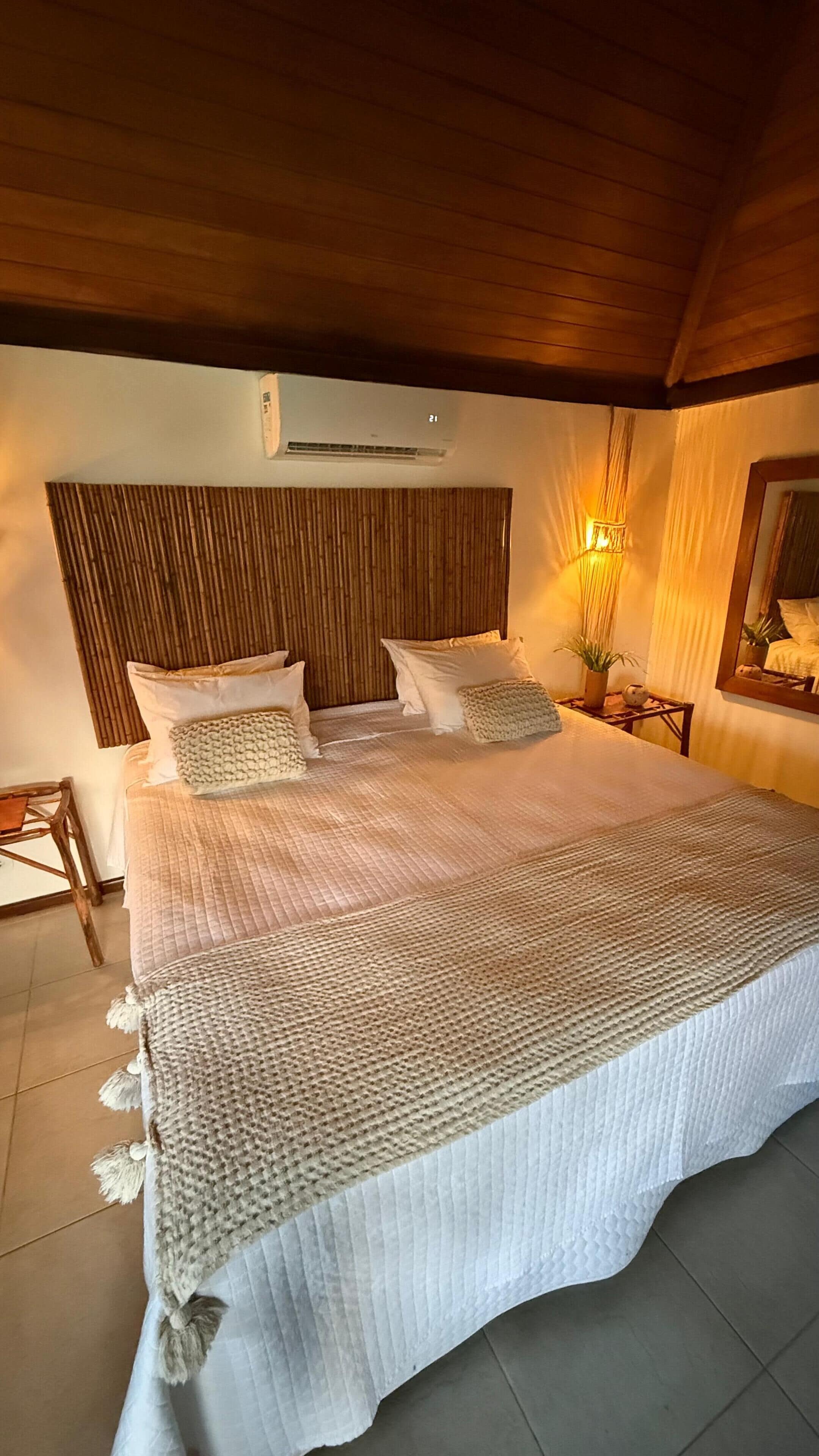 Una cama grande con cabecera de madera, una mesita de noche con lámpara y aire acondicionado de pared.