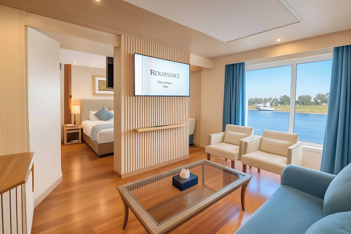 A modern hotel room with a bed, a seating area with a sofa and chairs, a TV mounted on the wall, and a large window with a view of the water.