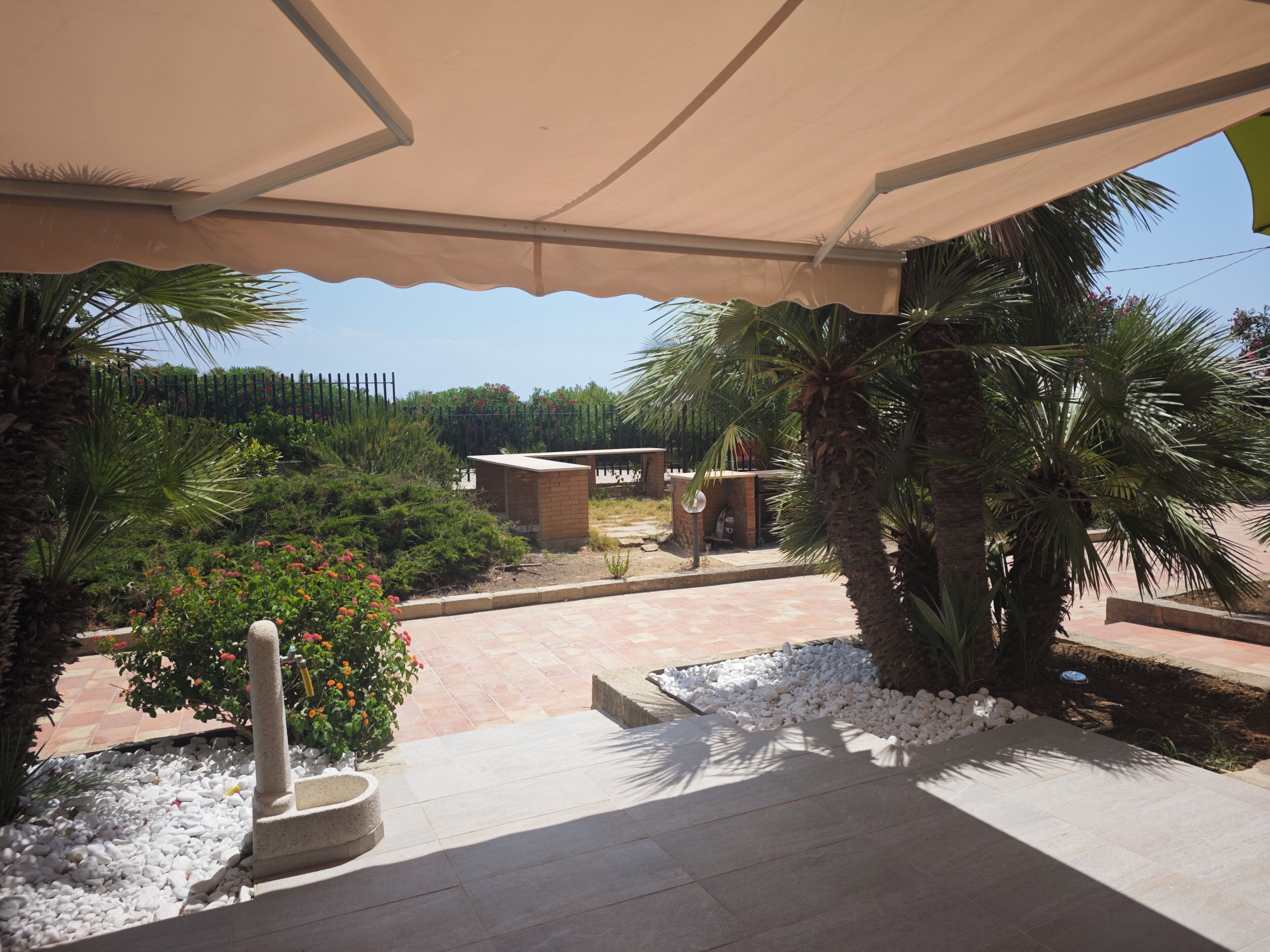 En udendørs patio med flisegulv, plantekasse i sten og en pergola der giver skygge.