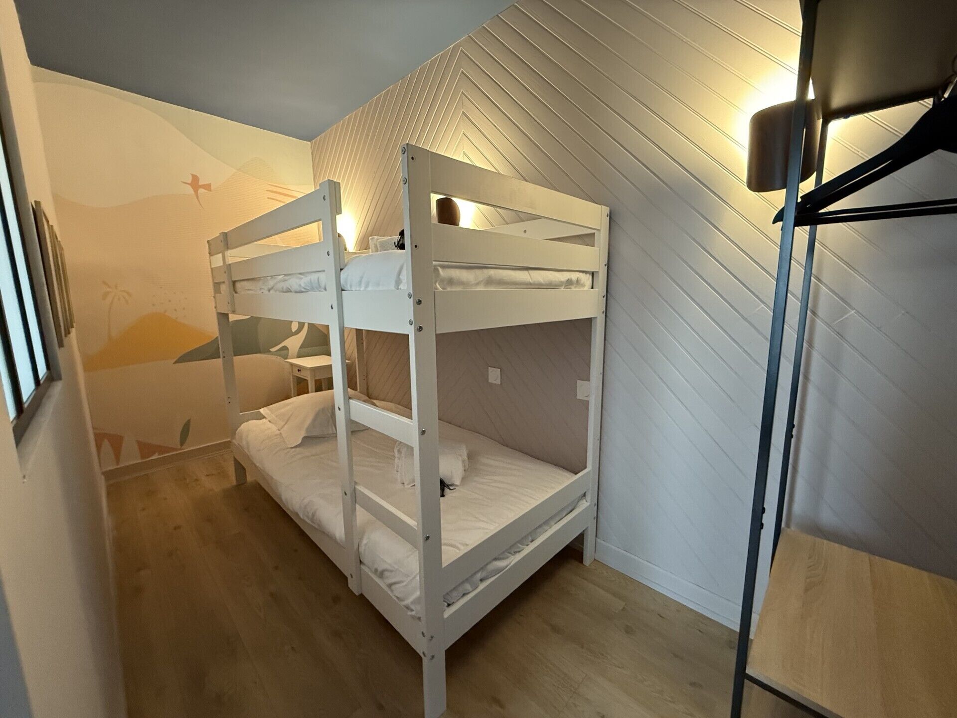 A bunk bed room with a wooden floor, a wall-mounted lamp, and a small table.