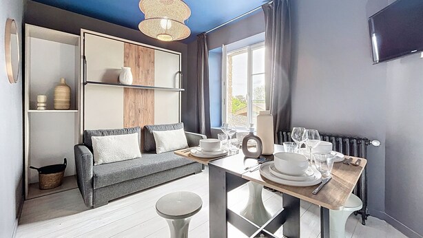 A modern living room with a sofa, a dining table set with plates and glasses, and a hanging light fixture. - Appart Hôtel de la Baie  (Saint-Jean-le-Thomas)