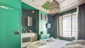 A modern hotel room with a bed, a kitchenette, a window with curtains, and a green accent wall. - Appart Hôtel de la Baie  (Saint-Jean-le-Thomas)