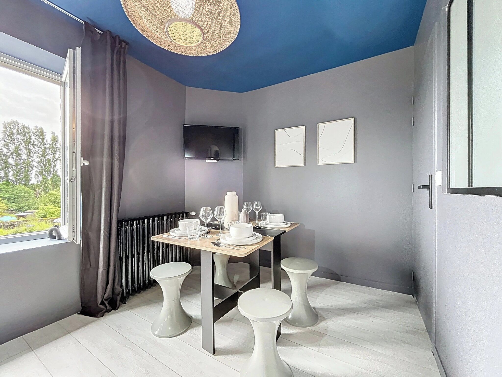 A modern dining area with a table set for four, a television on the wall, and a view of greenery outside.