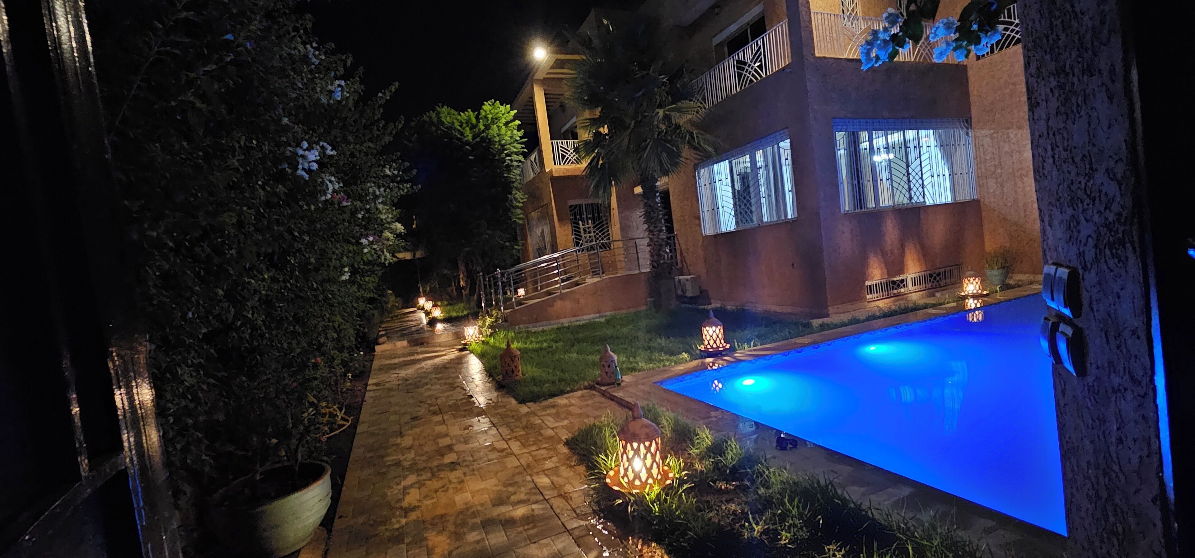 A nighttime view of a swimming pool area with a paved walkway, surrounded by greenery and illuminated by decorative lights.