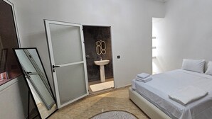 A bedroom with a bed, a nightstand, a mirror, a door, and a bathroom with a sink.
