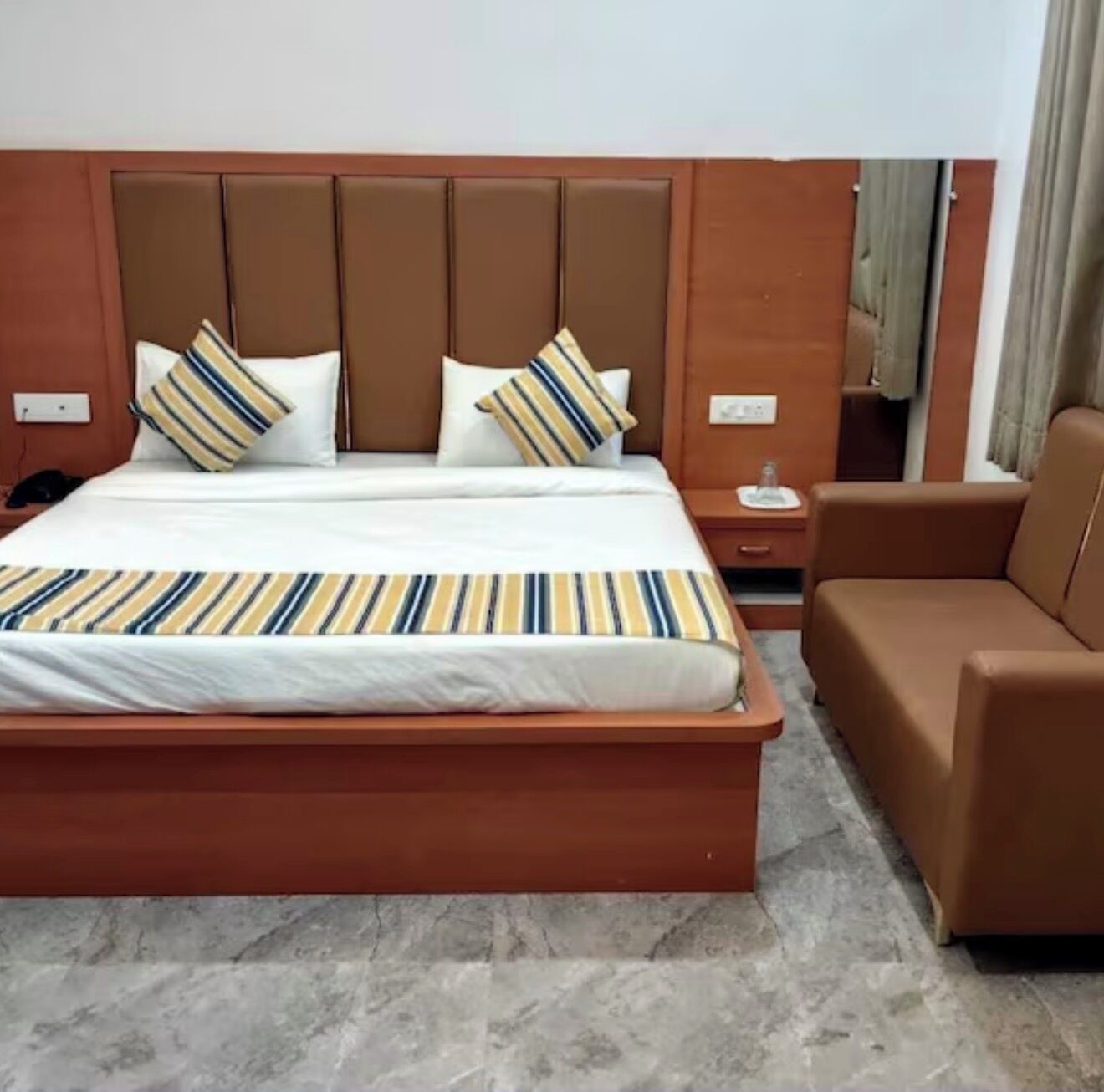 A hotel room with a bed, a nightstand, a bedside table, and a brown leather armchair.