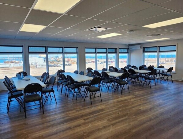 Un salón de conferencias con varias mesas y sillas, ventanas amplias con vistas a la playa y al puerto, y suelo de madera.