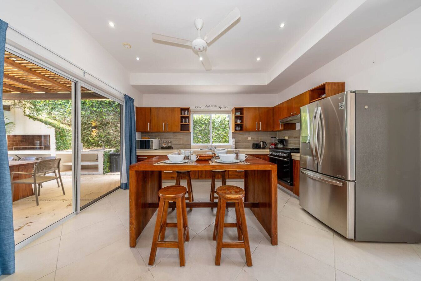 A modern kitchen with wooden cabinets, a stainless steel refrigerator, and a dining area with stools.