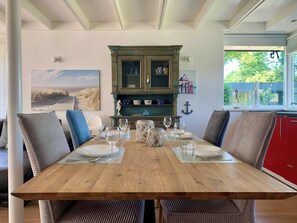 A dining area with a wooden table set for four, surrounded by upholstered chairs with tassels. A decorative cabinet with glass doors is against the wall, and a painting of a beach scene is visible. - Stylish Coastal Retreat in Sint Maartenszee (Sint Maartensvlotbrug)