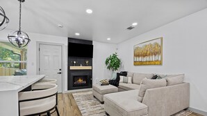 Un salon moderne avec un canapé beige, une table basse et une cheminée. On y trouve également un coin cuisine avec un comptoir blanc et des tabourets de bar. Un grand tableau est accroché au mur, et une plante est placée près de la cheminée.