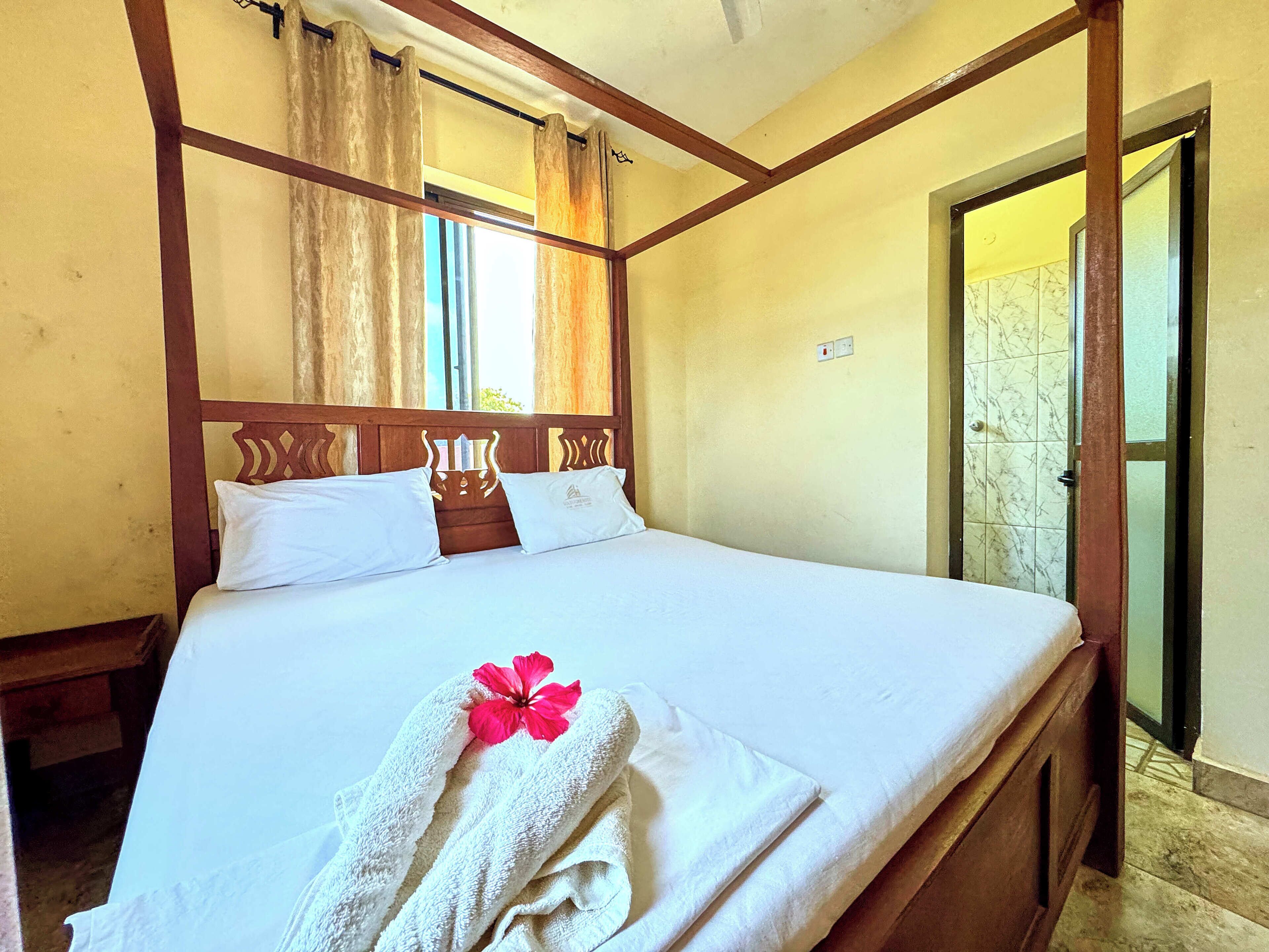Uma cama bem arrumada com roupa de cama branca e uma flor rosa, uma cabeceira de madeira e uma janela com cortinas.