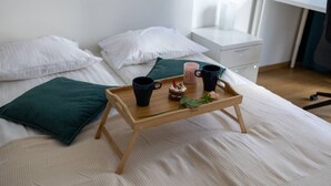 Een keurig opgemaakt bed met een houten dienblad waarop twee zwarte mokken, een roze mok en een klein taartje met een kaarsje staan.