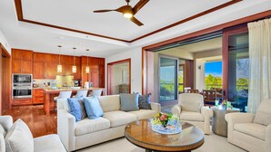 A modern living room with a sofa, coffee table, and chairs, a kitchen with wooden cabinets, and a large sliding glass door leading to a balcony.
