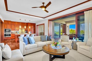 A modern living room with a sofa, coffee table, and chairs, a kitchen with wooden cabinets, and a large sliding glass door leading to a balcony.