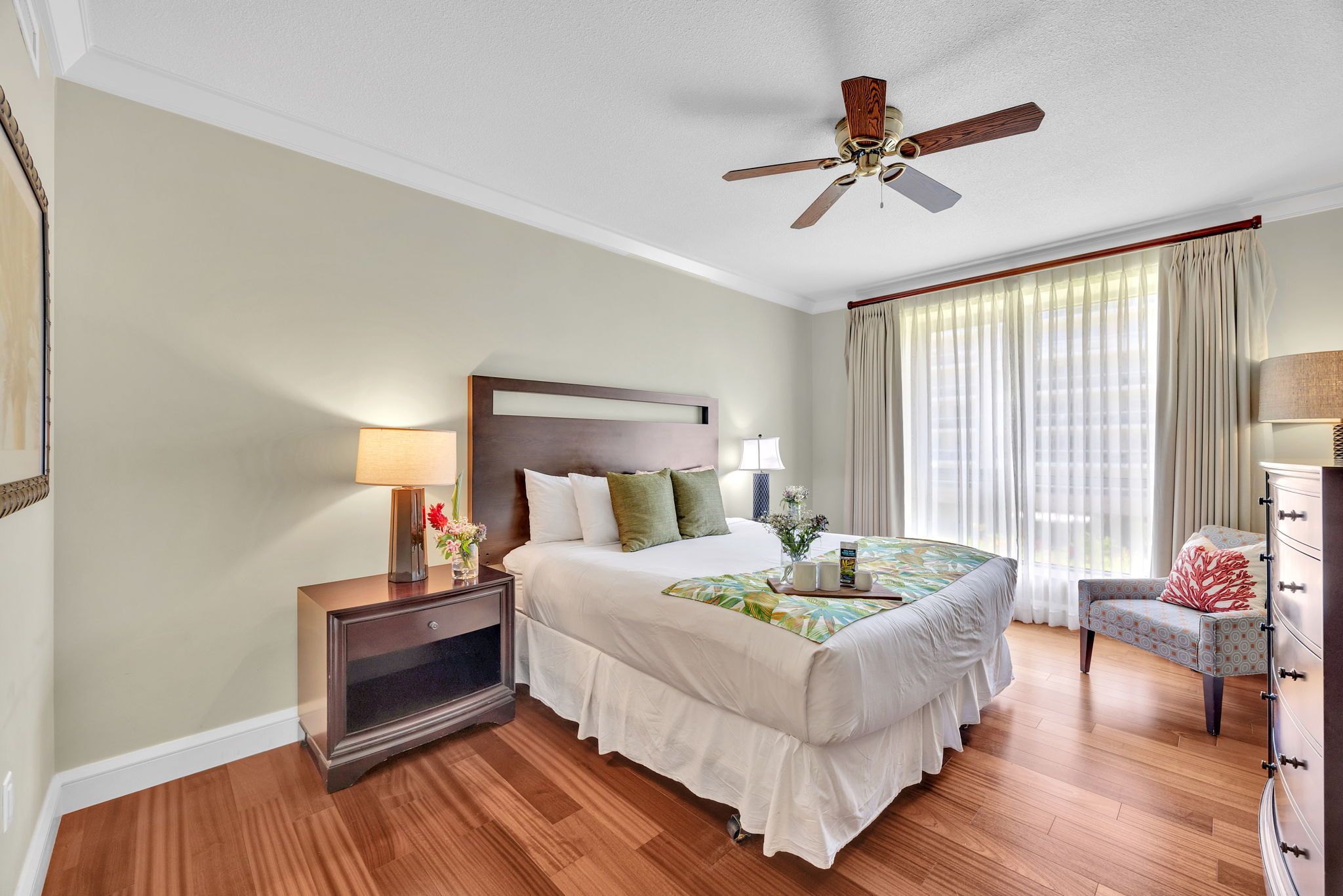 Une chambre d’hôtel avec un lit, une table de chevet, une chaise, un ventilateur de plafond et une grande fenêtre avec des rideaux.