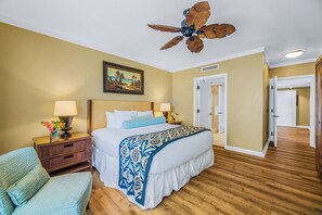 A hotel room with a bed, two bedside tables, a chair, a ceiling fan, a painting, and a door.