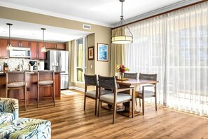 A modern kitchen with wooden cabinets, a dining area with a table and chairs, and a large window with sheer curtains.
