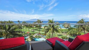 Un balcon agrémenté de meubles en osier et de coussins rouges, donnant sur une piscine et un paysage tropical avec des palmiers, et offrant une vue sur l’océan.
