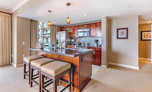 A modern kitchen with a central island, bar stools, wooden cabinets, stainless steel appliances, and pendant lighting.