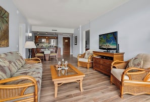 A living room with wicker furniture, a coffee table, a television, and a kitchen in the background.