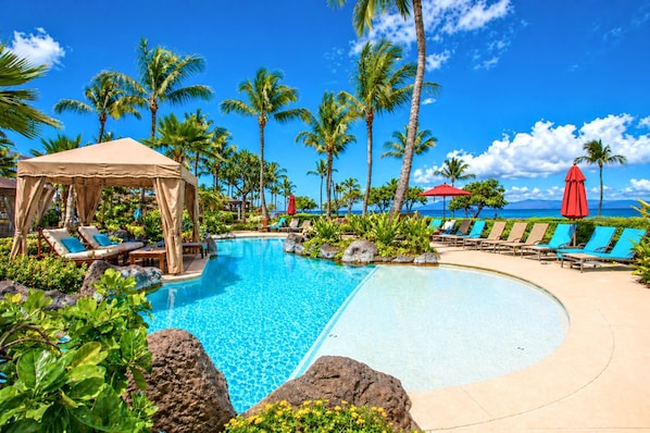A pool area with lounge chairs and a canopy, surrounded by palm trees and a clear sky.