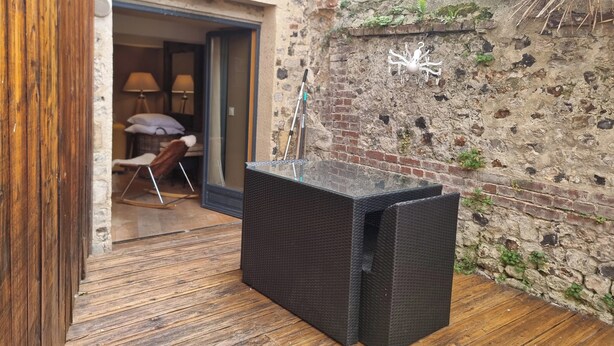 Un espacio exterior rústico con una silla de mimbre, una mesita y una puerta de madera que da a un interior con una lámpara y una cama.