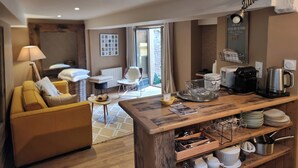 Habitación de hotel moderna con una mesa de comedor de madera, un sofá, una silla y una mesita auxiliar. Cuenta con una ventana con vistas al exterior, una lámpara y un estante con hervidor y cafetera.