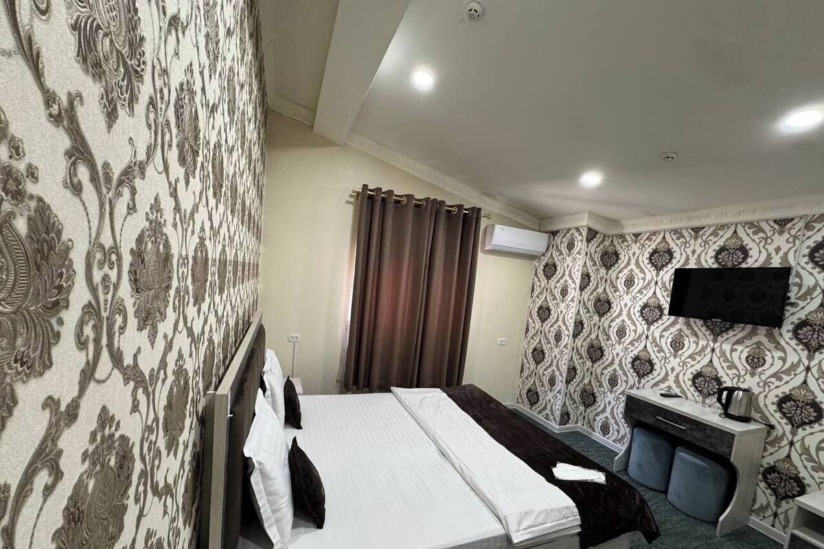 A hotel room with a bed, a desk with a chair, an air conditioning unit, and patterned wallpaper.