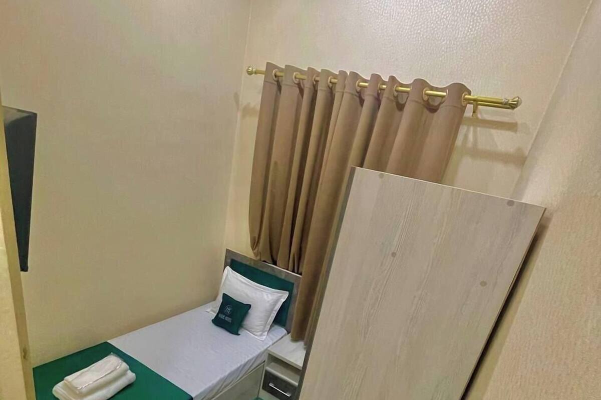 A single bed with a green mattress cover, a white pillow, and a beige curtain.