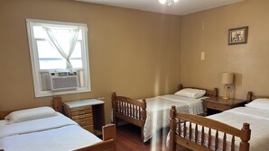 Ein Hotelzimmer mit drei hölzernen Betten, einem Holztisch, einem Fenster mit Vorhängen, einem Deckenventilator und einem gerahmten Bild an der Wand.