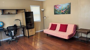 Ein Wohnzimmer mit Holzboden, einem pinken Sofa mit roten Kissen, einem schwarzen Bürostuhl, einem Schreibtisch mit Computer, einem Globus und einem Bild von Koi-Karpfen.