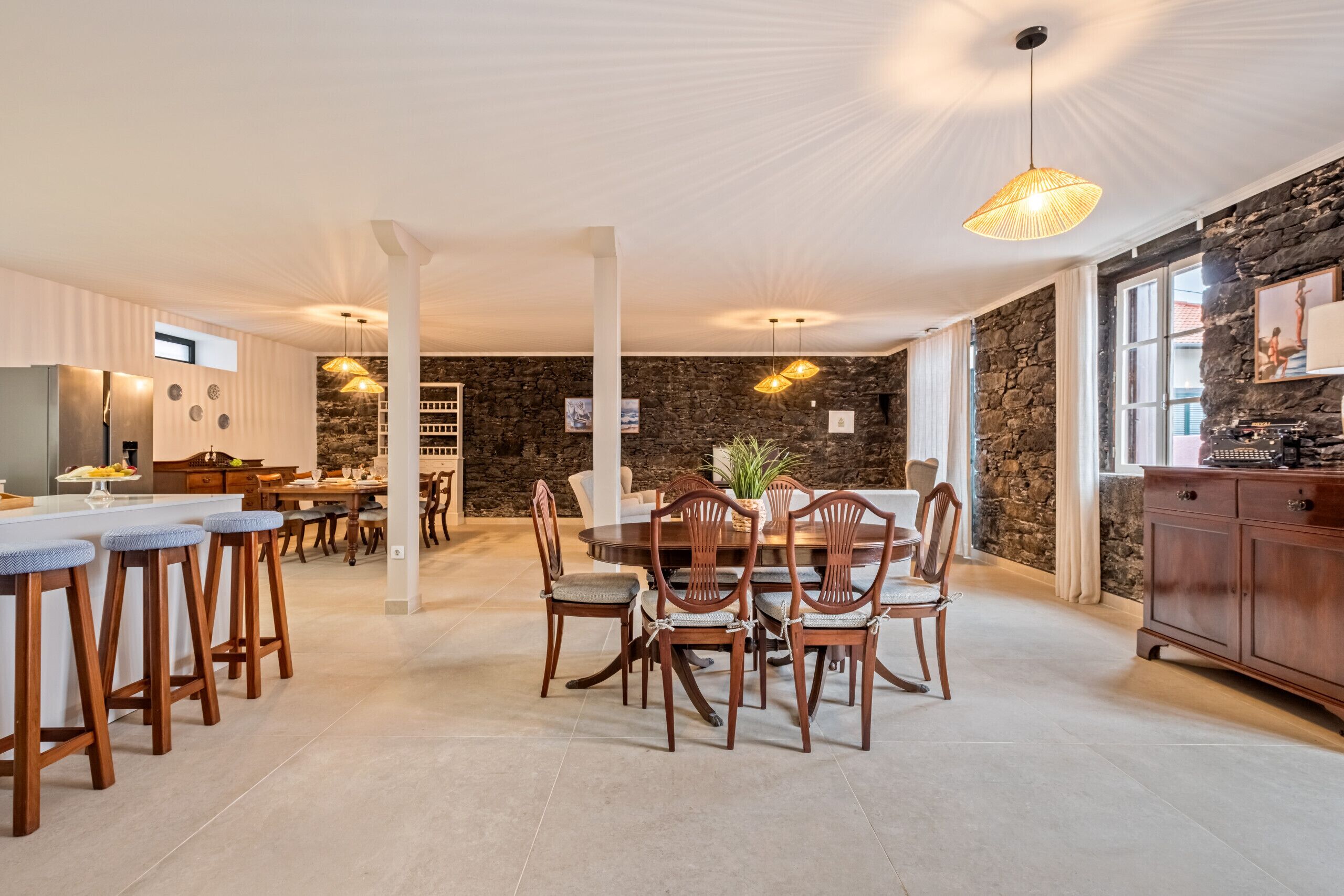 A spacious kitchen with a dining area, featuring a stone wall, wooden furniture, and a large window.