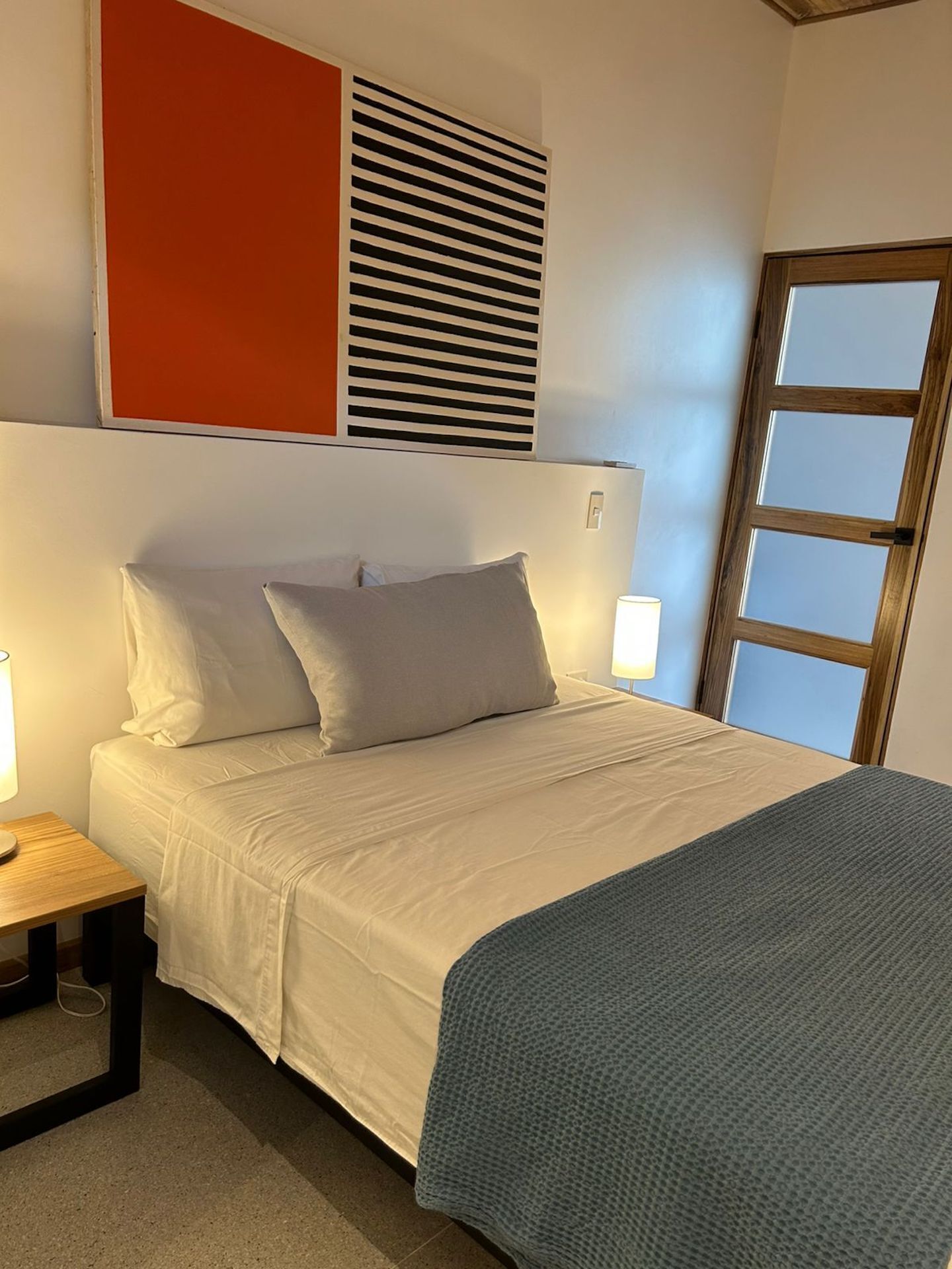 Een keurig opgemaakt bed met een wit-grijze dekbedovertrek, een blauwe deken en een houten nachtkastje met een lamp.