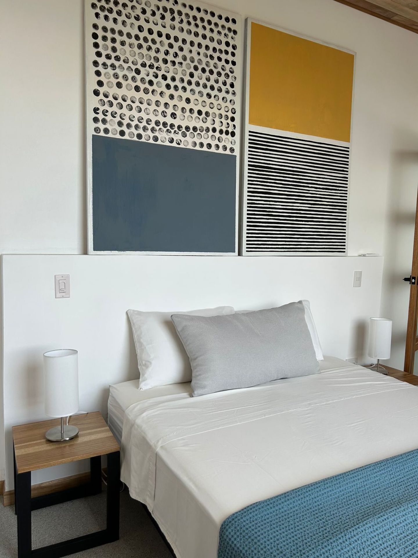 Une chambre à coucher avec un lit, deux oreillers, une table de chevet avec une lampe, et un mur orné de trois œuvres d’art.
