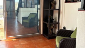 A room with a sliding glass door leading to an outdoor area, a wicker sofa with a green cushion, a black shelving unit with decorative items, and a white wall with a window. - Unwind at this peaceful oasis. 3 minute drive to the beach (Manzanillo)