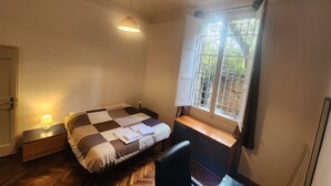 A small, well-lit bedroom with a checkered bedspread, a bedside table with a lamp, a window with a view of greenery, and a chair. - TATA HOMES B&B (Bologna)