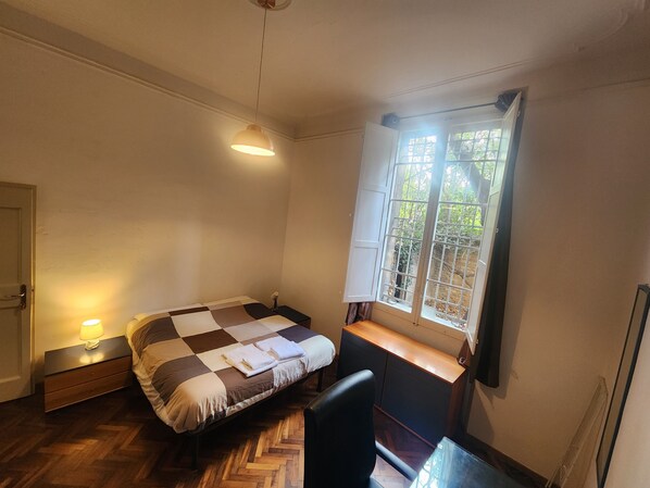 A small, well-lit bedroom with a checkered bedspread, a bedside table with a lamp, a window with a view of greenery, and a chair. - TATA HOMES B&B (Bologna)