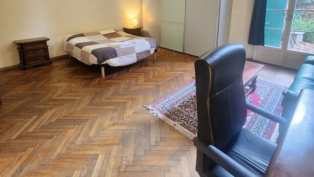 A room with a herringbone wooden floor, a bed with a striped bedspread, a black leather chair, a small table with a lamp, and a large window with a view of greenery outside. - TATA HOMES B&B (Bologna)