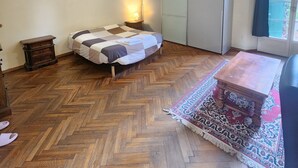A bedroom with a herringbone wooden floor, a bed with a checkered blanket, a nightstand, a wardrobe, and a window with a view. - TATA HOMES B&B (Bologna)